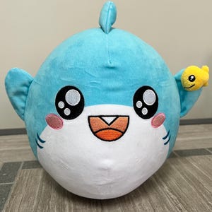 May include: A soft, round shark toy in light blue with a white belly. The shark has large, black eyes, pink cheeks, and a smiling orange mouth. A small yellow fish is attached to its fin. The toy is designed to be soft and cuddly.