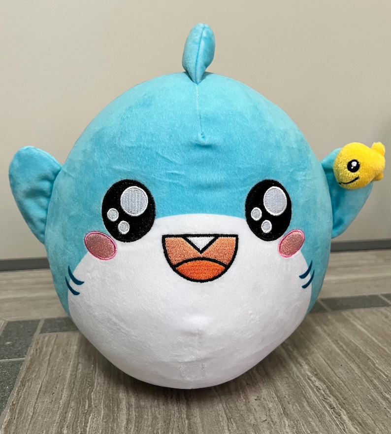 May include: A soft, round shark toy in light blue with a white belly. The shark has large, black eyes, pink cheeks, and a smiling orange mouth. A small yellow fish is attached to its fin. The toy is designed to be soft and cuddly.