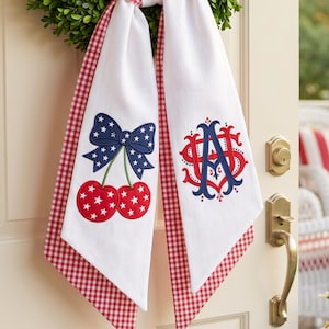 May include: A green boxwood wreath hangs on a door, adorned with a red and white gingham ribbon. Two white fabric tails hang down, one featuring an embroidered blue bow with white stars above two red cherries, and the other a red and blue monogram.