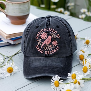 May include: A black baseball cap with the words "RADICALIZED BY BASIC DECENCY" in pink, along with a bird and floral design. The cap is on a light blue wooden surface, with a mug and books in the background.