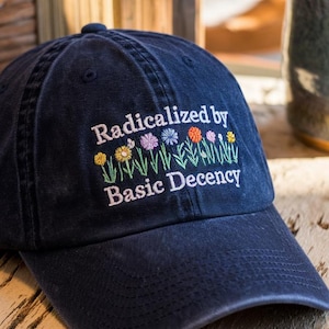 May include: A navy blue baseball cap with the text "Radicalized by Basic Decency" embroidered on the front. The text is surrounded by a row of colorful embroidered flowers.