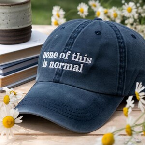 May include: A navy blue baseball cap with the embroidered text "none of this is normal" in white. The hat is on a wooden surface, surrounded by daisies and a ceramic mug.