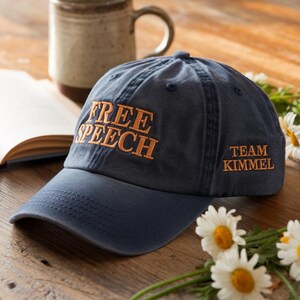 May include: A navy blue baseball cap with the words "FREE SPEECH" embroidered in orange on the front. The phrase "TEAM KIMMEL" is embroidered on the side. The hat sits on a wooden surface with a mug, book, and flowers.