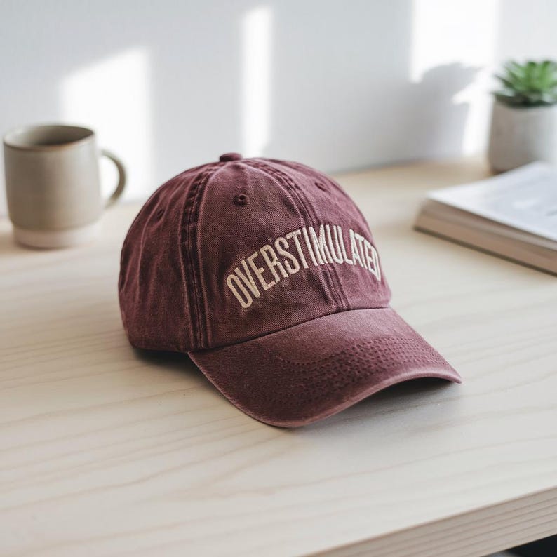 Peut inclure: Une casquette de baseball marron avec le texte "OVERStimulated" brod&eacute; en blanc sur le devant.