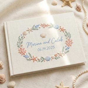 May include: A cream-colored fabric-covered book featuring an embroidered oval wreath of seashells, coral, and seaweed. The center is embroidered with the blue text Marina and Caleb and the date 06.14.2025 in a matching blue script font.