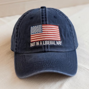 May include: A navy blue baseball cap with a curved brim. The cap features an embroidered American flag and the phrase "BUT IN A LIBERAL WAY" in white lettering. The cap is made of a soft, worn material.