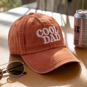 May include: A burnt orange baseball cap with the embroidered text "COOL DAD" in white. The cap is sitting on a wooden surface next to a can of beverage and a pair of sunglasses.
