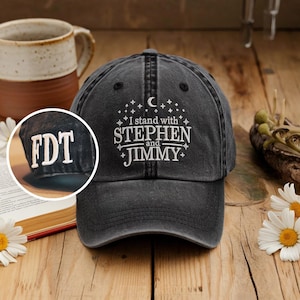 May include: A black, distressed baseball cap with white embroidered text that reads "I stand with Stephen and Jimmy." The cap also features the initials "FDT" embroidered on the side. The hat is sitting on a wooden surface with a mug and flowers in the background.