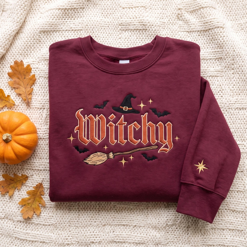 May include: A maroon crewneck sweatshirt featuring embroidered text that reads Witchy in a stylized font. The design includes a black witch hat, a broomstick, small black bats, and gold stars. An additional gold star is embroidered on the sleeve.