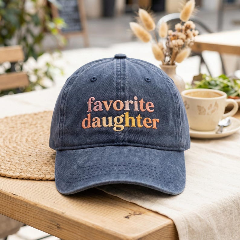 May include: A navy blue baseball cap with the embroidered text "favorite daughter" in a gradient of orange and yellow. The cap is on a wooden table with a woven placemat.