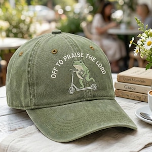 May include: Olive green baseball cap with the text "OFF TO PRAISE THE LORD" and a graphic of a frog riding a scooter while holding a book. The cap has a curved brim and a button on top.