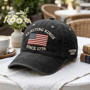 May include: A black baseball cap with the text "Rejecting Kings Since 1776" and an American flag design. The cap has a curved brim and a button on top. The words "Custom Text" are on the side.