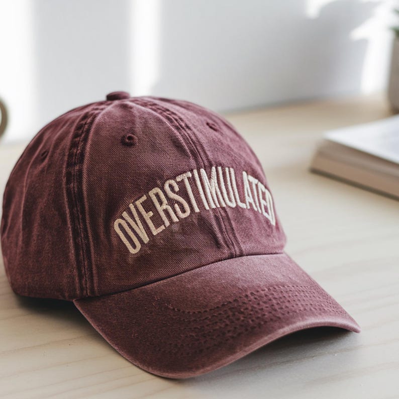 Peut inclure: Une casquette de baseball marron avec le texte "OVERStimulated" brod&eacute; en blanc sur le devant.