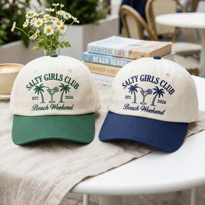 May include: Two baseball caps with the text "SALTY GIRLS CLUB" and "Beach Weekend". One cap has a green brim, the other has a navy blue brim. The caps are cream-colored with a palm tree and cocktail graphic.