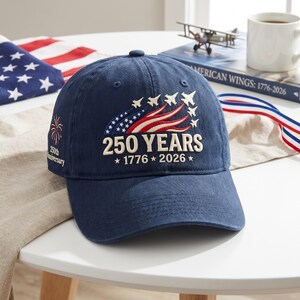 May include: A navy blue baseball cap featuring embroidered white text that reads 250 YEARS 1776 2026. The design includes a red, white, and blue flag graphic with white jet silhouettes and a firework icon with the text 250th Anniversary on the side.