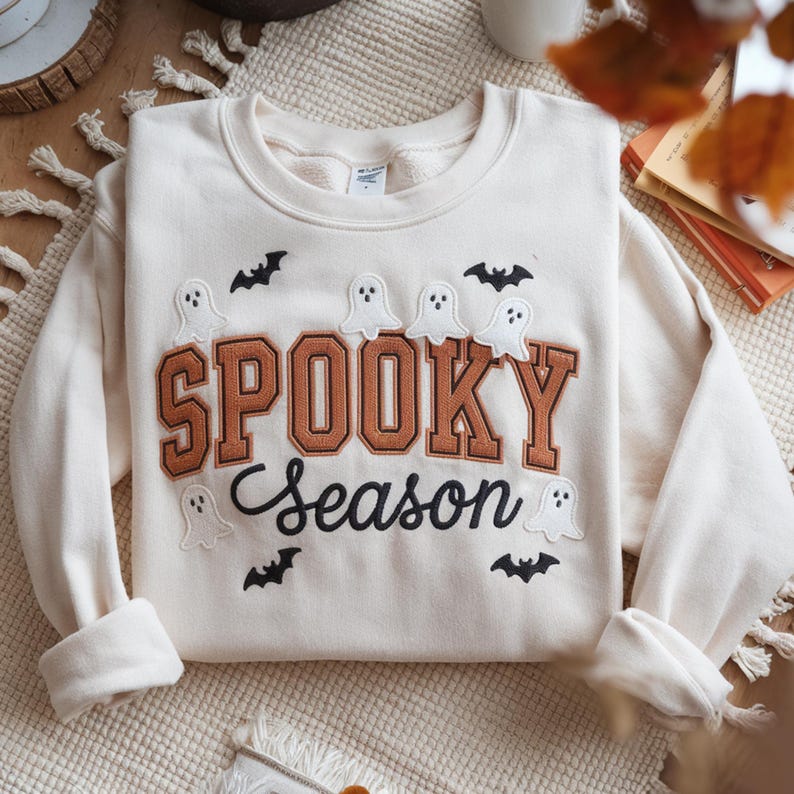 May include: Cream-colored sweatshirt with the words "SPOOKY Season" in brown and black lettering. The sweatshirt features embroidered ghosts and bats, perfect for Halloween or fall. The long-sleeved top is made of a soft material.