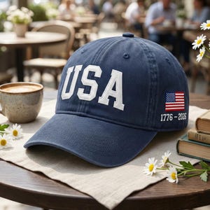 May include: A navy blue baseball cap with the word "USA" in white block letters. The cap features an American flag patch and the dates "1776-2026" on the side. The hat is sitting on a table with a cup of coffee and flowers.