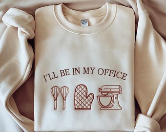 May include: Cream-colored sweatshirt with the embroidered text "I'LL BE IN MY OFFICE" above illustrations of a whisk, oven mitt, and stand mixer. The sweatshirt is folded and has a soft, casual appearance.