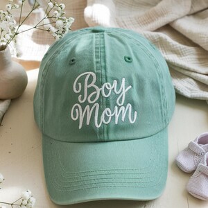 Embroidered green baseball cap with "Boy Mom" text in white thread, styled with baby shoes and soft beige fabrics, perfect gift for new moms of boys.