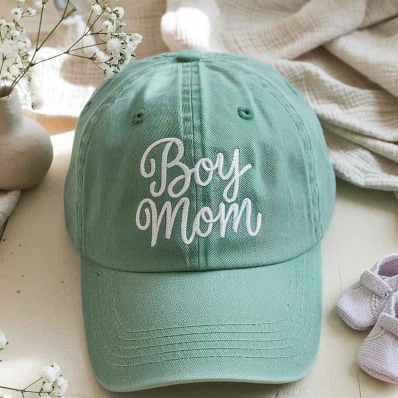 Embroidered green baseball cap with "Boy Mom" text in white thread, styled with baby shoes and soft beige fabrics, perfect gift for new moms of boys.