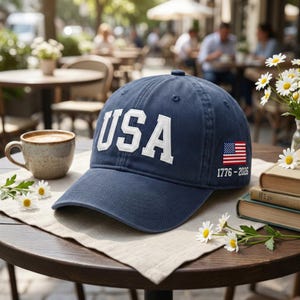 May include: A navy blue baseball cap with the word "USA" in white block letters. The cap features a small American flag and the dates "1776-2026" on the side. The cap is sitting on a table with a cup of coffee and flowers.