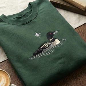 May include: A dark green sweatshirt featuring an embroidered design of a loon swimming in water, with a white star to the left. The loon has black, white, and gray feathers, a red eye, and a black head.