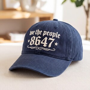 May include: Navy blue baseball cap with the text "we the people 8647" in white script. The cap has a curved brim and a fabric strap closure. The hat is made of a soft, durable material.
