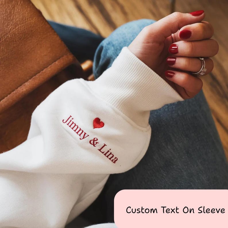 May include: White sweatshirt sleeve with the embroidered names "Jimmy & Lina" in red, along with a small red heart. The sleeve is ribbed at the cuff. The image also shows a brown bag and blue jeans.