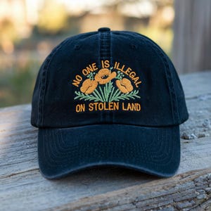 May include: Black baseball cap with embroidered orange flowers and green leaves. The text "NO ONE IS ILLEGAL ON STOLEN LAND" is embroidered in orange. The cap is made of a soft, durable fabric.