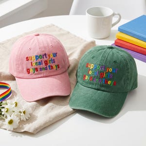 May include: A pink baseball cap and a green baseball cap, both featuring the embroidered text 'support your local girls gays and theys' in multi-colored letters. Each cap has a curved brim and a structured crown with ventilation eyelets.