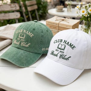 May include: Two baseball caps, one green and one white, with embroidered text. The green cap has "CLUB NAME EST. 2025 Book Club" in white, and "02.08". The white cap has the same text in green.