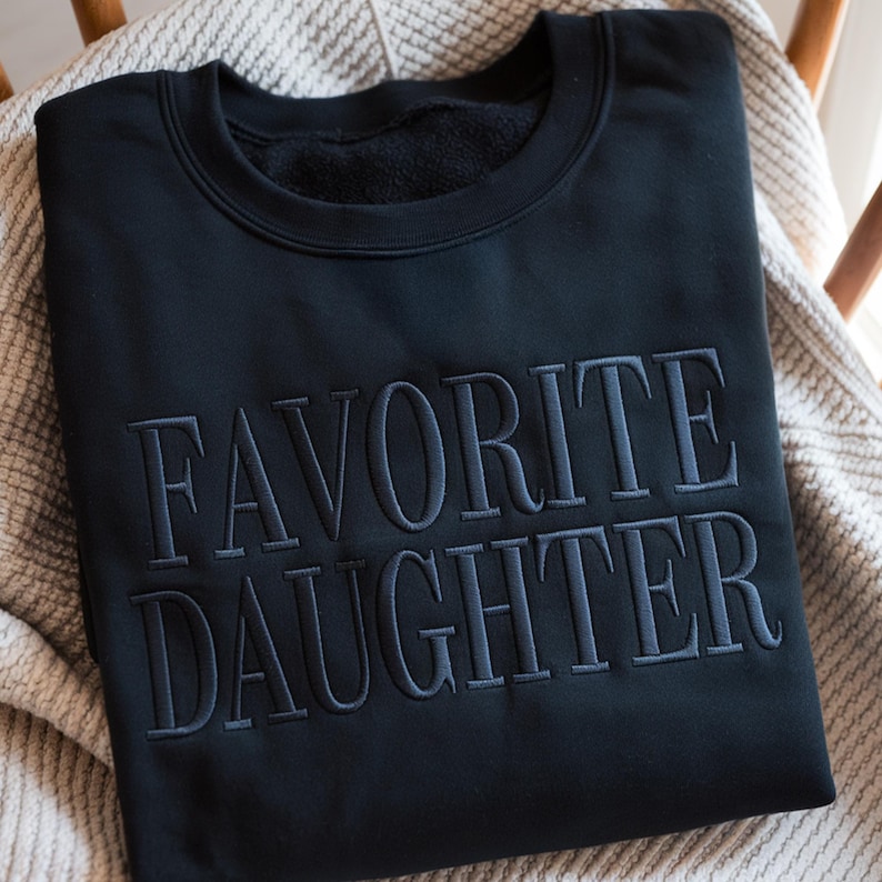May include: Black sweatshirt with the embroidered text "FAVORITE DAUGHTER" in a darker shade of gray. The sweatshirt is folded and resting on a light-colored, textured blanket. The crew neck sweatshirt is made of a soft material.