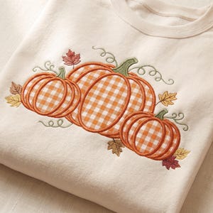 May include: A cream-colored crewneck sweatshirt featuring an embroidered design of three pumpkins. The pumpkins have an orange and white gingham pattern with green stems and vines, surrounded by small embroidered autumn leaves in brown and gold.