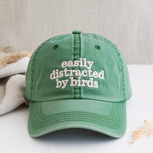 May include: A green baseball cap with the embroidered text "easily distracted by birds" in white. The cap has a curved brim and a button on top. The hat is made of a soft, worn material.