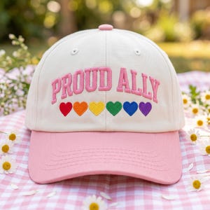 May include: A baseball cap with a cream-colored crown and a pink bill. The cap features the embroidered text "PROUD ALLY" in pink, above a row of rainbow-colored hearts. The cap is a stylish accessory.