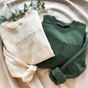Pode incluir: Duas camisolas de gola redonda, uma creme e outra verde escuro, são exibidas sobre um tecido neutro. A palavra "YWHW" é bordada em uma cor correspondente em cada camisola. As mangas estão dobradas e o tecido tem uma aparência macia e texturizada.