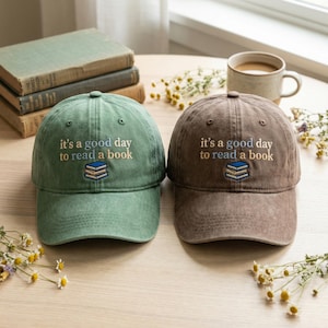 May include: Two baseball caps, one sage green and one brown, sit on a wooden table. Each cap has the embroidered phrase "it's a good day to read a book" above a stack of books. A cup of coffee and old books are in the background.