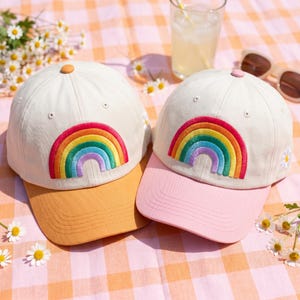 May include: Two baseball caps with embroidered rainbow designs. One cap has a tan brim and the other has a pink brim. Both caps are off-white with a small daisy embroidered on the side. The caps are on a checkered picnic blanket.