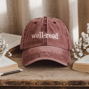 May include: A maroon baseball cap with the embroidered text "well-read" in white. The hat is sitting on a wooden surface next to open books and white flowers. The cap has a curved brim and a classic six-panel design.