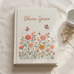 May include: A cream-colored fabric-covered book featuring the embroidered name Olivia Grace in brown script. The cover displays a colorful botanical scene with various flowers, green stems, two butterflies, and a small bee stitched in thread.