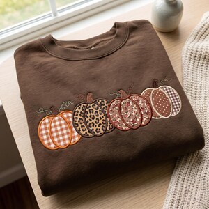 May include: A dark brown crewneck sweatshirt featuring an embroidered row of four pumpkins. Each pumpkin displays a different pattern: orange and white gingham, brown leopard print, a floral print, and brown and white polka dots.
