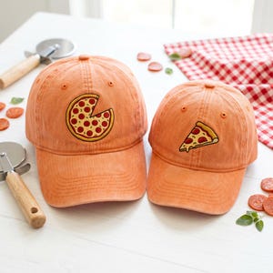 May include: Two orange baseball caps with embroidered pizza slice designs. One cap features a pizza slice, and the other has a pizza with a slice missing. The caps are on a white surface with pizza cutters and pepperoni slices.