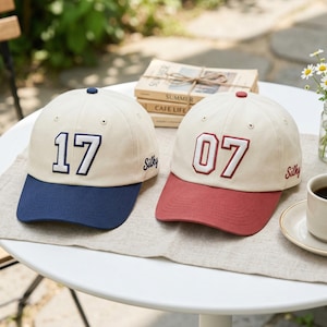 May include: Two baseball caps with cream-colored crowns and colored brims. One has a navy blue brim and the number "17" embroidered in navy. The other has a red brim and the number "07" embroidered in red. Both caps have the word "Silky" embroidered on the side.