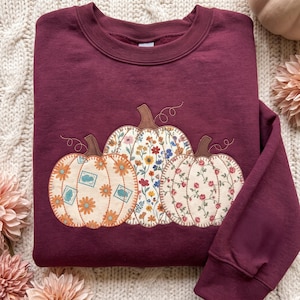 May include: A deep maroon crewneck sweatshirt featuring an embroidered applique design of three pumpkins. Each pumpkin displays a distinct floral pattern in shades of orange, blue, yellow, and pink, with brown stems and decorative stitching.