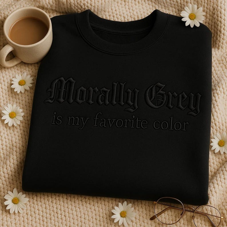 Puede incluir: Sudadera negra con la frase bordada "Morally Grey is my favorite color". La imagen incluye una taza de caf&eacute;, gafas y margaritas.