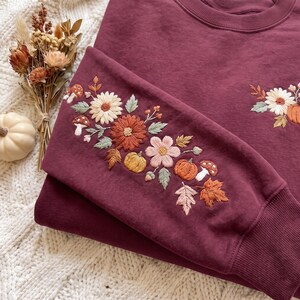 K&ouml;nnte beinhalten: Ein tiefburgunderfarbenes Sweatshirt mit Rundhalsausschnitt und aufwendigen Stickereien an &Auml;rmel und Brust. Das Design zeigt cremefarbene, orange und braune Blumen, kleine K&uuml;rbisse, Pilze und Herbstbl&auml;tter, die direkt in den Stoff gestickt sind.