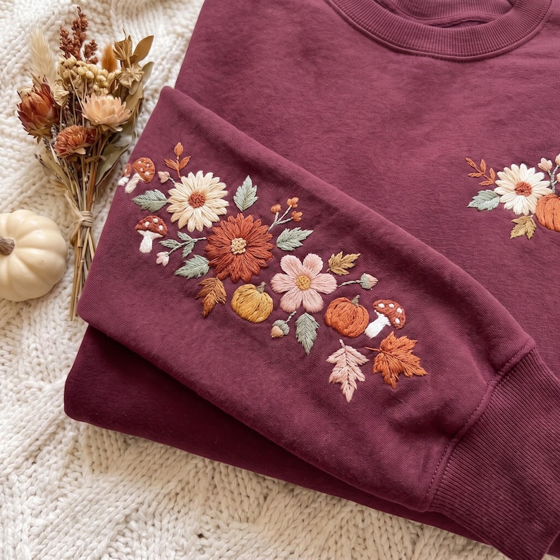 K&ouml;nnte beinhalten: Ein tiefburgunderfarbenes Sweatshirt mit Rundhalsausschnitt und aufwendigen Stickereien an &Auml;rmel und Brust. Das Design zeigt cremefarbene, orange und braune Blumen, kleine K&uuml;rbisse, Pilze und Herbstbl&auml;tter, die direkt in den Stoff gestickt sind.