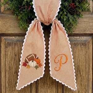 May include: A decorative fabric bow featuring a brown and white vertical stripe pattern with white scalloped edges. One tail displays an embroidered football, two pumpkins, and autumn leaves, while the other tail features a large orange letter P.