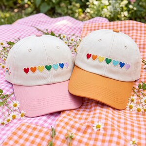 May include: Two baseball caps with cream-colored crowns and colorful heart embroidery. One has a pink bill, the other an orange bill. A rainbow is embroidered on the side of the pink-billed cap. The caps are on a checkered blanket.