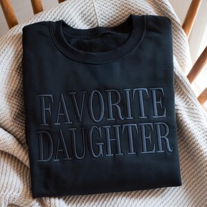 May include: Black sweatshirt with the embroidered text "FAVORITE DAUGHTER" in a darker shade of gray. The sweatshirt is folded and resting on a light-colored blanket, placed on a wooden chair.